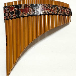 May include: A wooden panpipe with a black and red patterned band. The panpipe is made of natural bamboo and has a curved shape.