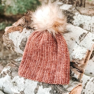 May include: A coral-coloured knitted beanie hat with a large, fluffy, light brown faux fur pom-pom. The hat is displayed on a stack of birch logs, with a blurred natural background.