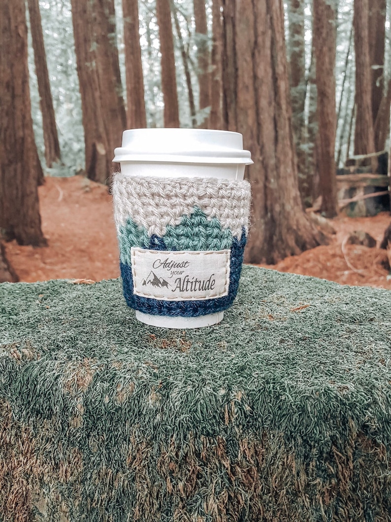 May include: A white coffee cup with a crocheted cozy in shades of beige, blue, and green. The cozy has the text "Adjust your Altitude" on it.