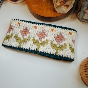 Può includere: Un cerchietto all'uncinetto con sfondo bianco e un motivo di foglie verdi e fiori rosa. Il cerchietto è rifinito con un bordo verde scuro.