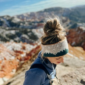 Può includere: Una donna con un chignon disordinato indossa una fascia per capelli all'uncinetto con strisce bianche, beige e verdi. La fascia è indossata sulla fronte e copre le orecchie. Indossa una giacca di pile blu e guarda in basso.