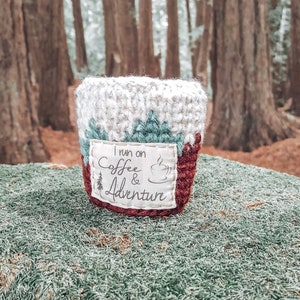 May include: A crocheted coffee cup cozy with a red, white, and green design. The cozy has a white patch with the text "I run on coffee & adventure" and a small coffee cup icon.