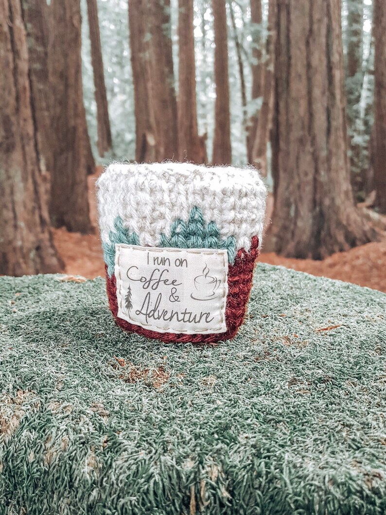 May include: A crocheted coffee cup cozy with a red, white, and green design. The cozy has a white patch with the text "I run on coffee & adventure" and a small coffee cup icon.