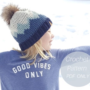 Op de afbeelding: Een haakpatroon voor een blauwe, grijze en witte beanie met een nep bont pompon. Het patroon is alleen beschikbaar als PDF-download. De tekst "Good Vibes Only" is gedrukt op een blauw shirt.