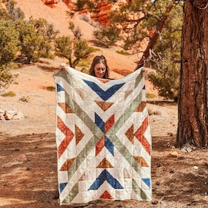 May include: A person is holding up a large crocheted blanket with a geometric pattern in shades of blue, green, orange, and white. The blanket is displayed in front of a red rock landscape.
