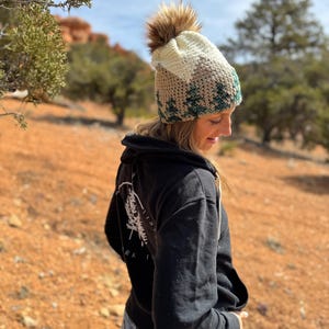 Op de afbeelding: Een vrouw draagt een zwarte hoodie met een witte afbeelding van een dennenboom op de rug. Ze draagt een gebreide muts met een bruin, wit en groen kleurpatroon en een bruine nep bontpompon bovenop.