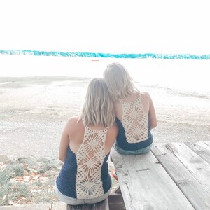 May include: Two people wearing navy blue tank tops with crocheted white floral designs on the back, sitting on a wooden bench facing a body of water.