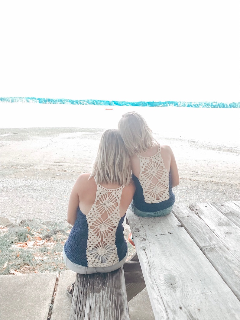 May include: Two people wearing navy blue tank tops with crocheted white floral designs on the back, sitting on a wooden bench facing a body of water.