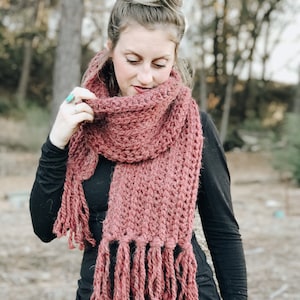 May include: A long, burgundy crocheted scarf with fringe. The scarf is worn by a person with their hair in a bun.