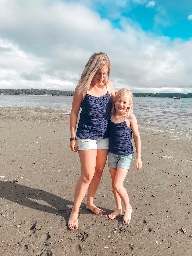 May include: Two people, an adult and a child, wearing matching navy blue crochet tank tops. They are standing on a sandy beach with a blue sky and white clouds in the background.