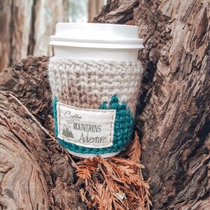 May include: A crocheted coffee cup cozy with a beige and teal design. The cozy has a white fabric patch with the text "Coffee Mountains Adventure".