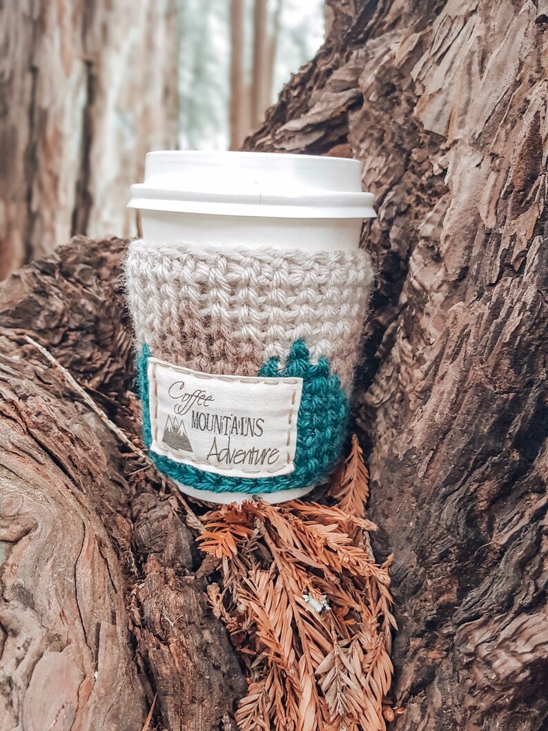 May include: A crocheted coffee cup cozy with a beige and teal design. The cozy has a white fabric patch with the text "Coffee Mountains Adventure".