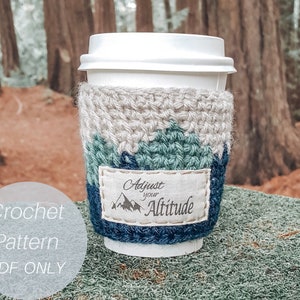 May include: Crochet pattern for a coffee cup cozy with a mountain design and the text "Adjust your Altitude".