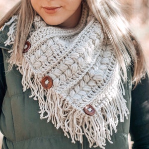 May include: A crocheted cowl with a textured stitch pattern and fringe. The cowl has three wooden buttons.