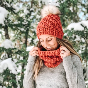 May include: A woman wearing a crocheted beanie and matching scarf in a burnt orange color. The beanie has a fluffy white pom pom on top. The woman is smiling and looking down.