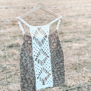 May include: A brown crochet tank top with a white crochet panel down the back. The tank top is hanging on a wooden hanger against a backdrop of a field.