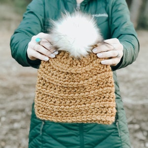 May include: A brown crocheted beanie with a white faux fur pom pom. The beanie is being held by a person wearing a green jacket.