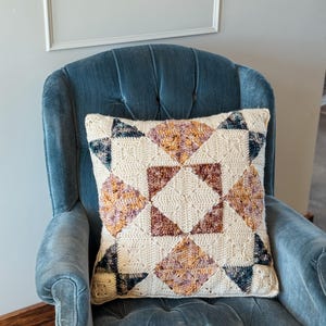 May include: A cream-colored, square crochet pillow with a geometric design. The pattern features diamond shapes in shades of brown, orange, purple, and navy blue. The pillow is placed on a blue velvet armchair.