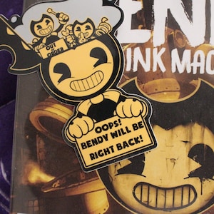 Puede incluir: Un personaje de dibujos animados negro y amarillo, Bendy, del videojuego "Bendy and the Ink Machine", sostiene un cartel que dice "¡Ups! ¡Bendy volverá enseguida!" El cartel es amarillo con letras negras. El fondo es una portada de videojuego con un personaje de dibujos animados negro y amarillo.
