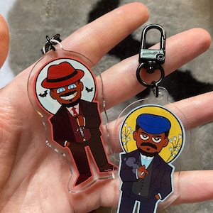 May include: Two clear acrylic keychains featuring cartoon character illustrations. One shows a figure in a red hat and suit holding a knife against a moon background. The other depicts a figure in a blue cap and suit smoking, with a yellow halo background.