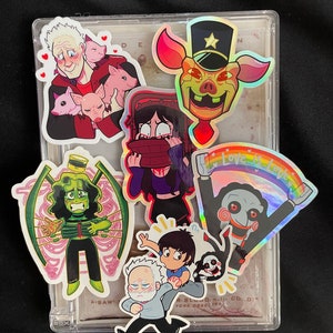 May include: A collection of colourful stickers featuring characters from popular horror movies. The stickers are displayed in a clear plastic case. The stickers include a pig wearing a hat, a girl with a red mask, a man with a chainsaw, a man with a pig mask, and a man with a saw. The stickers are all holographic and have a rainbow effect.