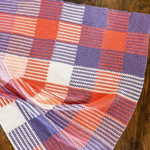 May include: A crocheted blanket with a checkered pattern in shades of purple, pink, white, and orange.