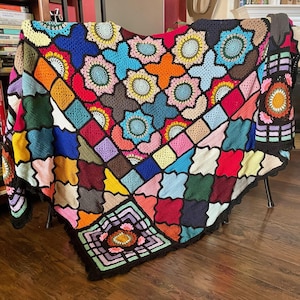 May include: A colorful, handmade crochet blanket with a patchwork design. The blanket features a variety of geometric shapes and floral motifs in shades of red, blue, green, yellow, and pink. The edges are finished with a black scalloped border.