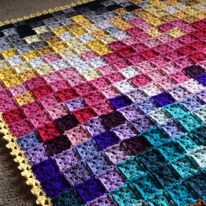 Op de afbeelding: Een handgemaakte gehaakte deken met een patchwork-ontwerp. De deken heeft vierkanten in tinten zwart, grijs, geel, roze, rood, paars en turquoise. De randen zijn afgewerkt met een gele geschulpte rand.