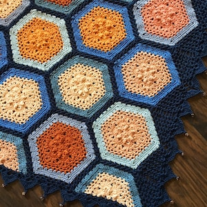May include: A crocheted blanket with a hexagonal pattern in shades of blue, orange, and beige. The blanket has a dark blue border with a beaded fringe.