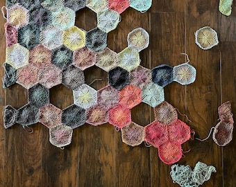 May include: A crocheted blanket made of colorful hexagonal patches in shades of pink, red, blue, green, and brown. The blanket is laid out on a wooden surface and some of the patches are not yet attached.
