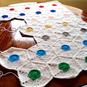 May include: A white crocheted blanket with a geometric pattern of hexagons. Each hexagon has a different colored circle in the center, including blue, green, yellow, orange, red, and gray.