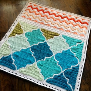 May include: A crocheted blanket with a geometric pattern in shades of blue, green, and white. The blanket has a white border and a chevron pattern in shades of orange and pink at the top.