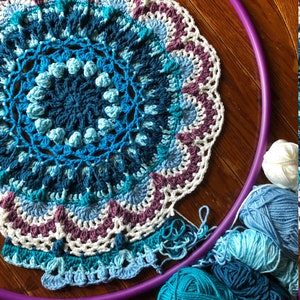 Op de afbeelding: Een gehaakt mandala met een paarse hoepel en blauw, wit en paars garen. De mandala is gemaakt met verschillende steken en heeft een gegolfde rand.