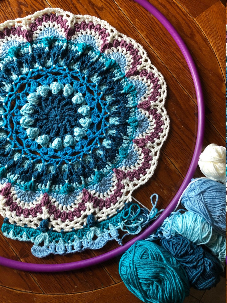 Op de afbeelding: Een gehaakt mandala met een paarse hoepel en blauw, wit en paars garen. De mandala is gemaakt met verschillende steken en heeft een gegolfde rand.