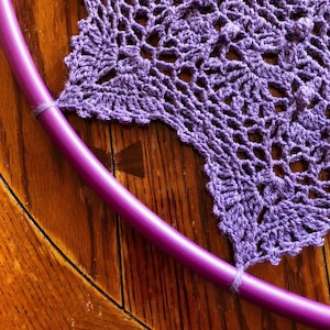 Op de afbeelding: Een paarse gehaakte doily met een paarse hoepel die op een houten oppervlak ligt.