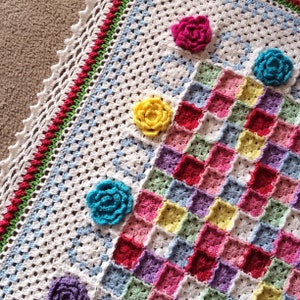 May include: A white crochet blanket with a colorful patchwork design. The blanket features a border of red, green, and white crocheted flowers. The blanket is made with a variety of colors, including pink, blue, yellow, and green.
