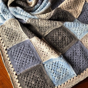 May include: A crocheted baby blanket with a patchwork design in shades of blue and gray. The blanket has a white crocheted border.