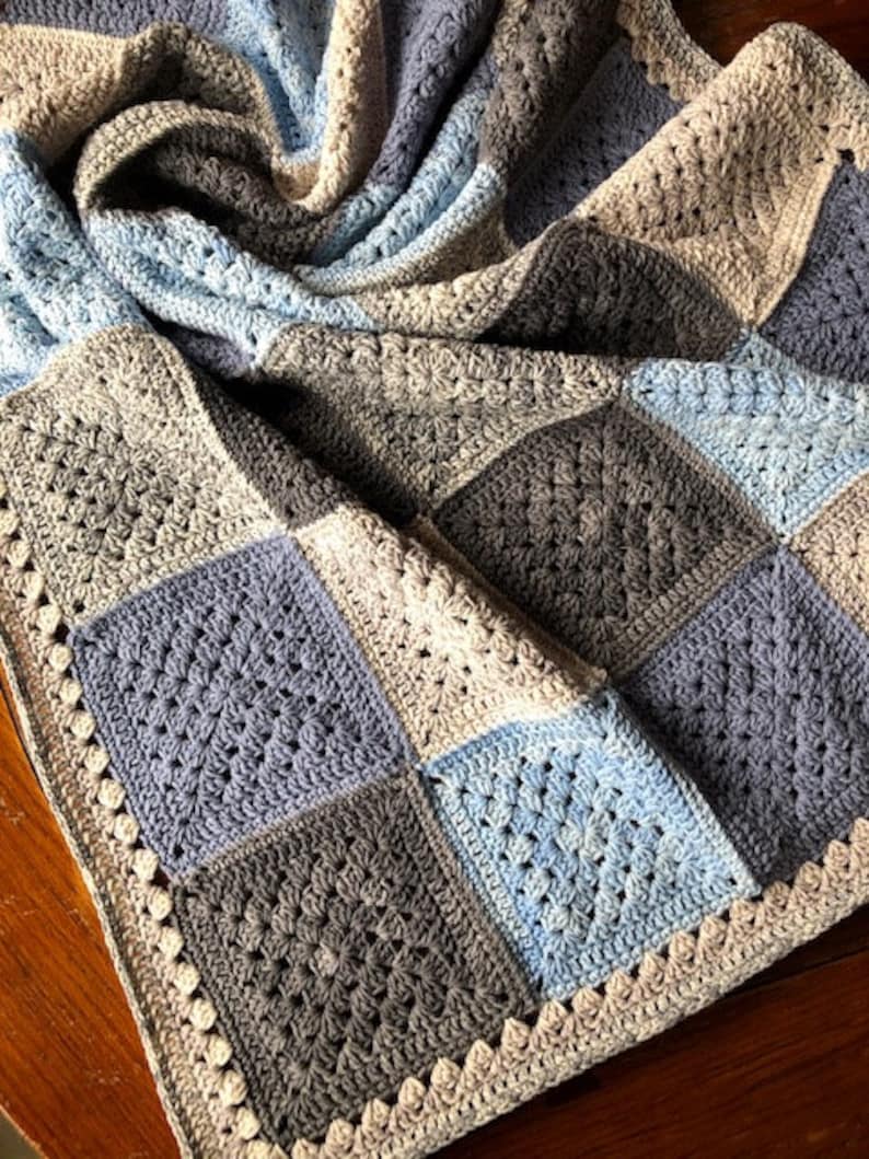 May include: A crocheted baby blanket with a patchwork design in shades of blue and gray. The blanket has a white crocheted border.