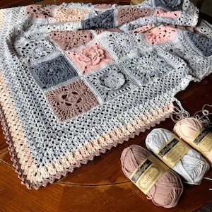 May include: A crocheted baby blanket in shades of gray, pink, and beige with a crocheted flower detail. Three skeins of yarn in pink, gray, and beige are in the foreground. The yarn labels read "Scheepjes Softfun Double Knit."