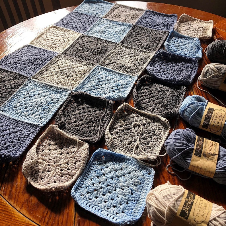 May include: A crocheted blanket in shades of blue, gray, and beige, with several skeins of yarn in matching colors. The blanket is made of squares, each with a different pattern. The yarn is labeled "Softies" and "Softies Denim".