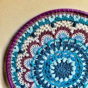 Op de afbeelding: Een grote, ronde, gehaakte wandhanger met een paarse rand. Het midden van de hanger is een mandala-ontwerp met blauw, turquoise en wit garen.