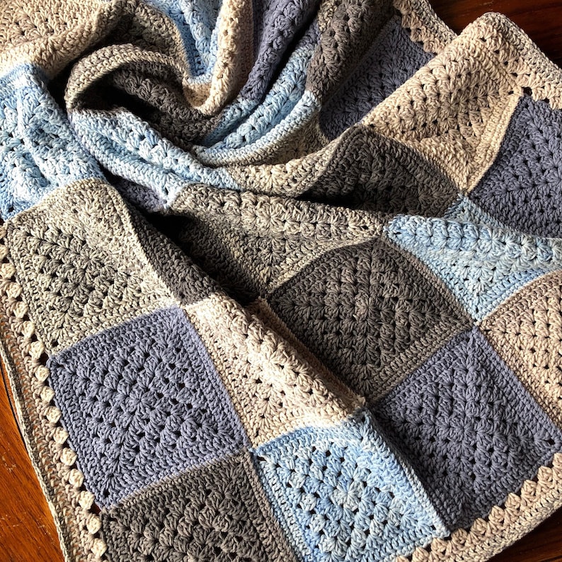 May include: A crocheted blanket with a square pattern in shades of blue, gray, and beige. The blanket is made with a variety of stitches and has a textured look.