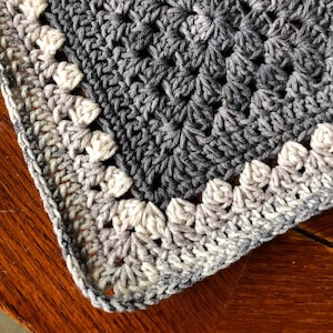 May include: A crocheted blanket with a gray and white ombre border. The blanket is made with a granny square stitch and has a textured, woven look.