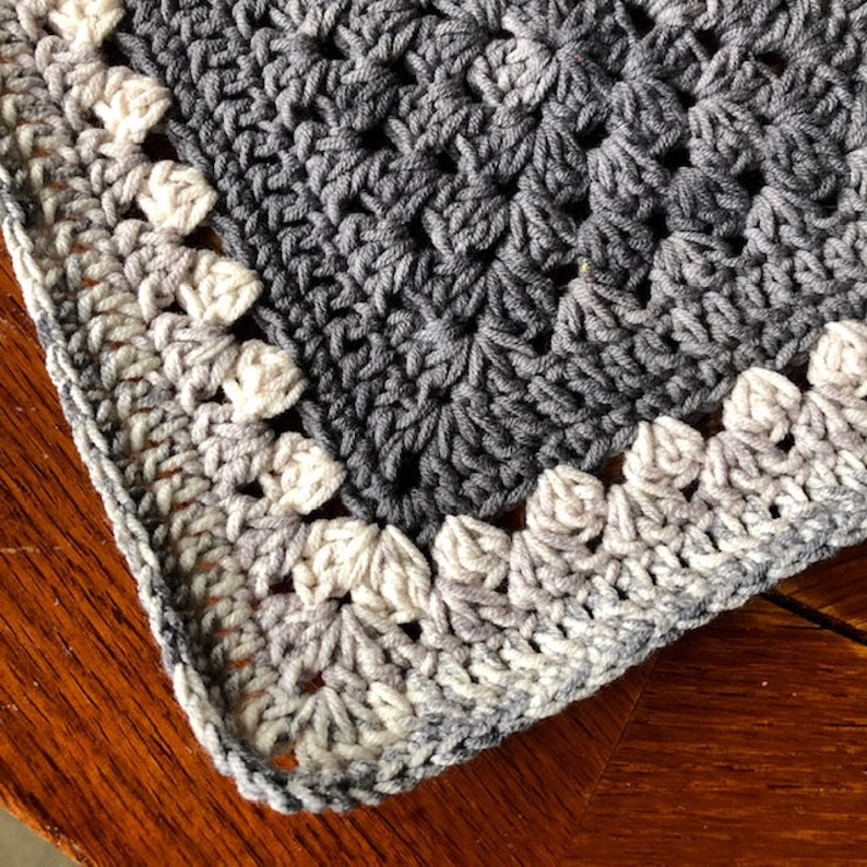 May include: A crocheted blanket with a gray and white ombre border. The blanket is made with a granny square stitch and has a textured, woven look.