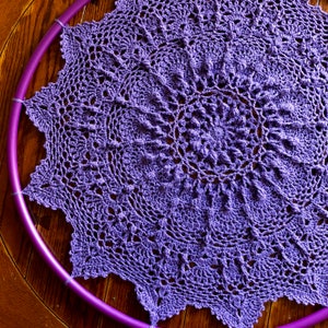 Op de afbeelding: Een paarse gehaakte doily met een cirkelvormig ontwerp. De doily ligt plat op een houten oppervlak.