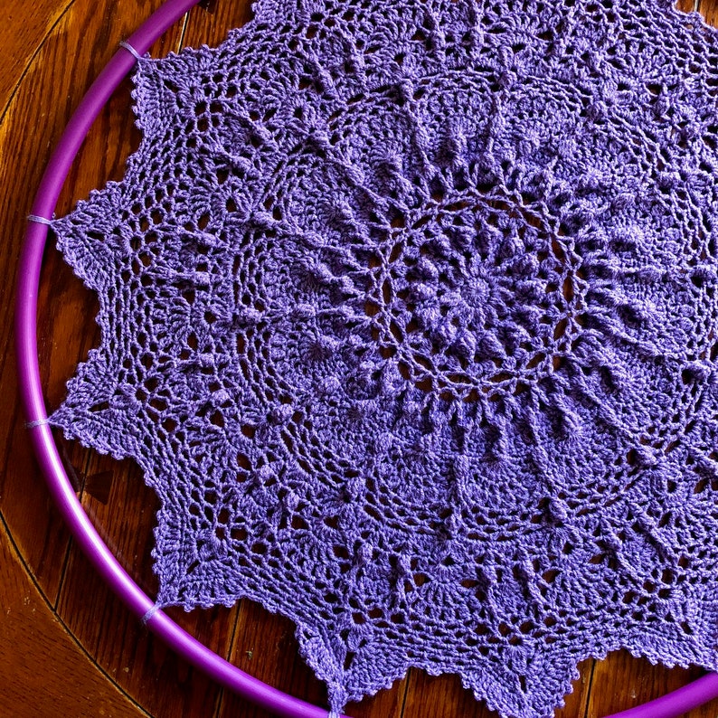 Op de afbeelding: Een paarse gehaakte doily met een cirkelvormig ontwerp. De doily ligt plat op een houten oppervlak.