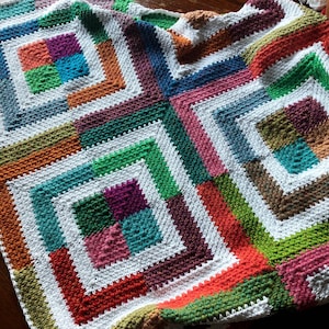 May include: A colourful crocheted blanket with a geometric pattern of squares within squares. The blanket is made with a variety of colours, including red, orange, yellow, green, blue, purple, and white.