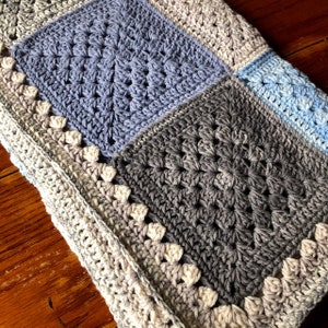 May include: A crocheted baby blanket with a patchwork design in shades of blue and gray. The blanket has a white border with a scalloped edge.