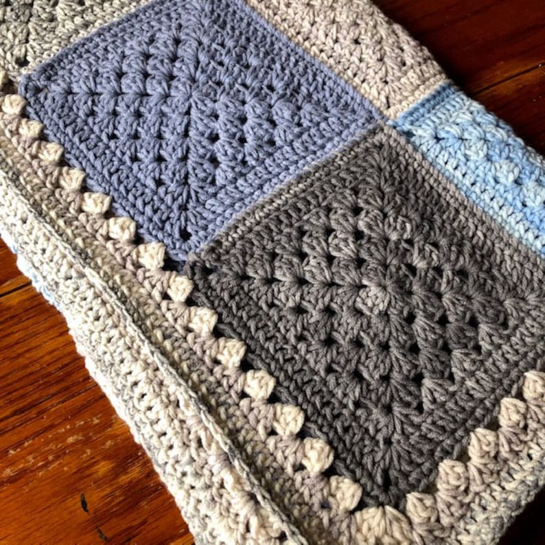 May include: A crocheted baby blanket with a patchwork design in shades of blue and gray. The blanket has a white border with a scalloped edge.