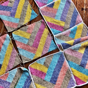 May include: A collection of colourful crocheted squares with a chevron pattern in shades of pink, blue, yellow, and brown. The squares are arranged in a grid pattern and are joined together with white yarn.